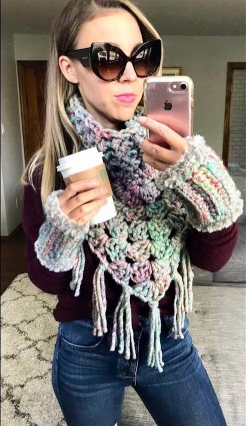 May include: A woman wearing a multi-coloured crochet scarf and matching arm warmers with long tassels. She is also wearing a maroon sweater and blue jeans.