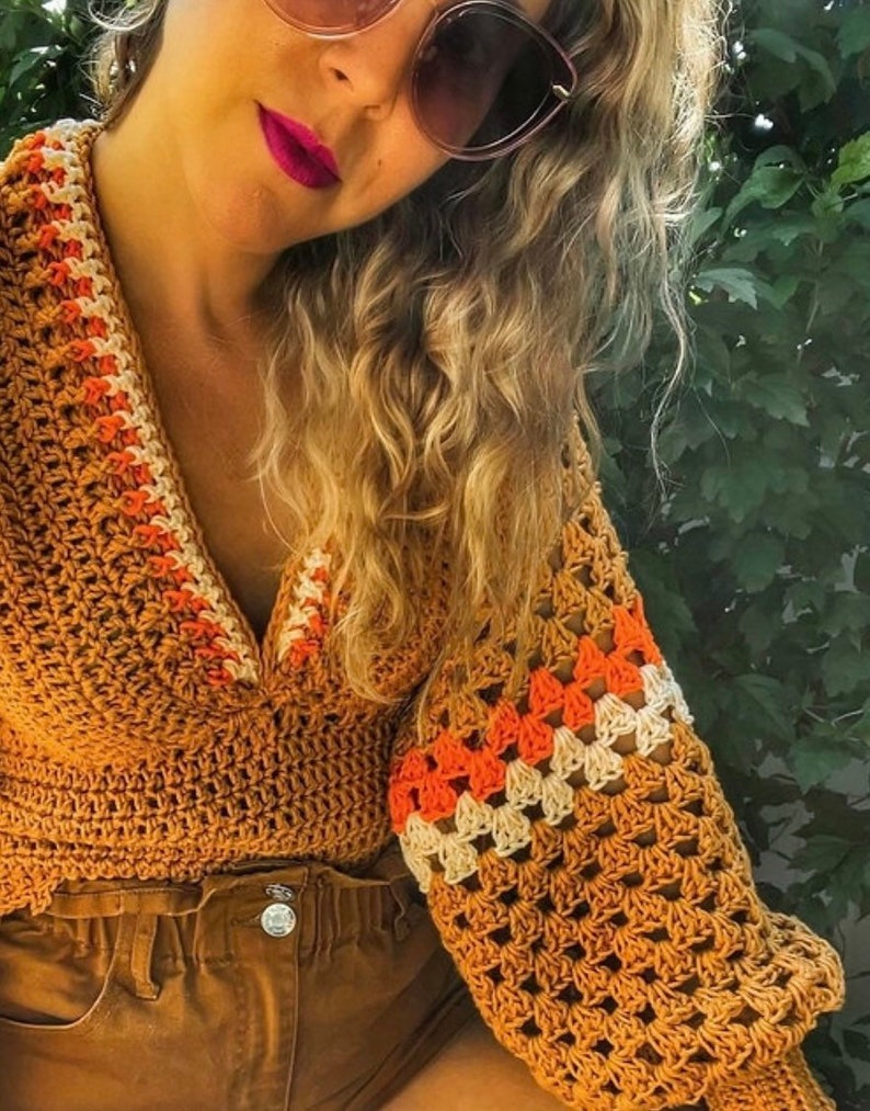 May include: A crocheted top with a V-neck and long sleeves. The top is made in a brown color with orange and white stripes on the sleeves. The top is cropped and has a loose fit.