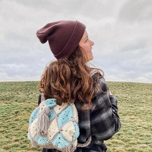 May include: A woman wearing a brown beanie and a plaid shirt is carrying a crocheted backpack with a grey tassel. The backpack is made of white, blue, and grey yarn and has a diamond pattern.