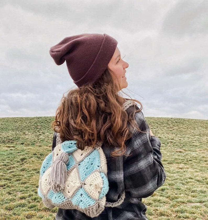 May include: A woman wearing a brown beanie and a plaid shirt is carrying a crocheted backpack with a grey tassel. The backpack is made of white, blue, and grey yarn and has a diamond pattern.