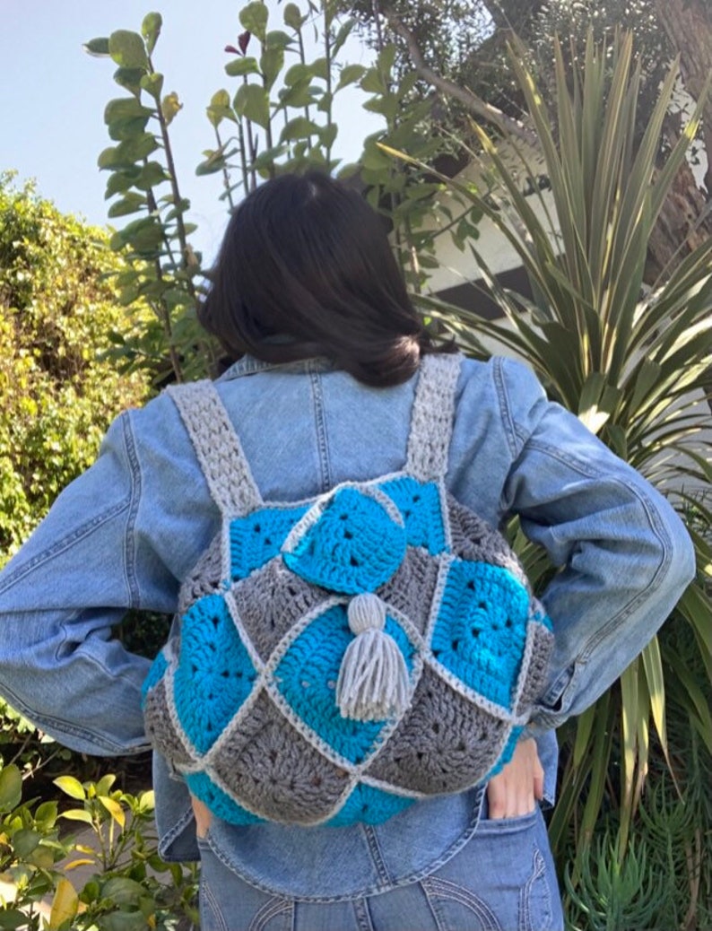 May include: A crocheted backpack with a grey and blue diamond pattern. The backpack has a grey tassel.