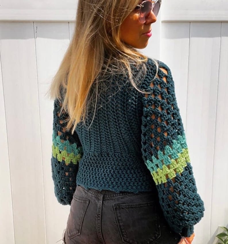 May include: A teal blue crocheted sweater with green and light blue stripes on the sleeves. The sweater has a loose fit and a crochet stitch pattern.