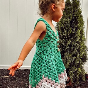 May include: A green crochet dress with a scalloped hemline in white and pink. The dress features a button-up front and shoulder straps. The dress is handmade and suitable for children.