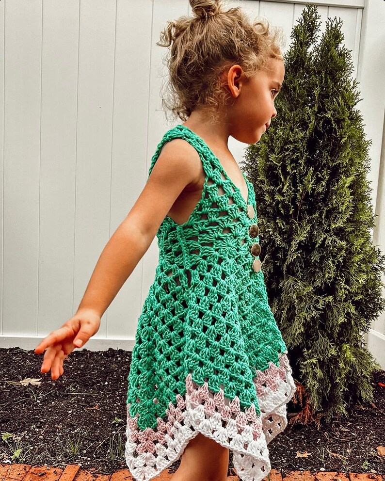 May include: A green crochet dress with a scalloped hemline in white and pink. The dress features a button-up front and shoulder straps. The dress is handmade and suitable for children.