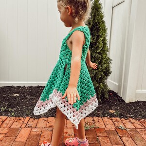 May include: A green crochet dress with a white and pink border. The dress is sleeveless and has a flared skirt. The child is wearing pink clogs.