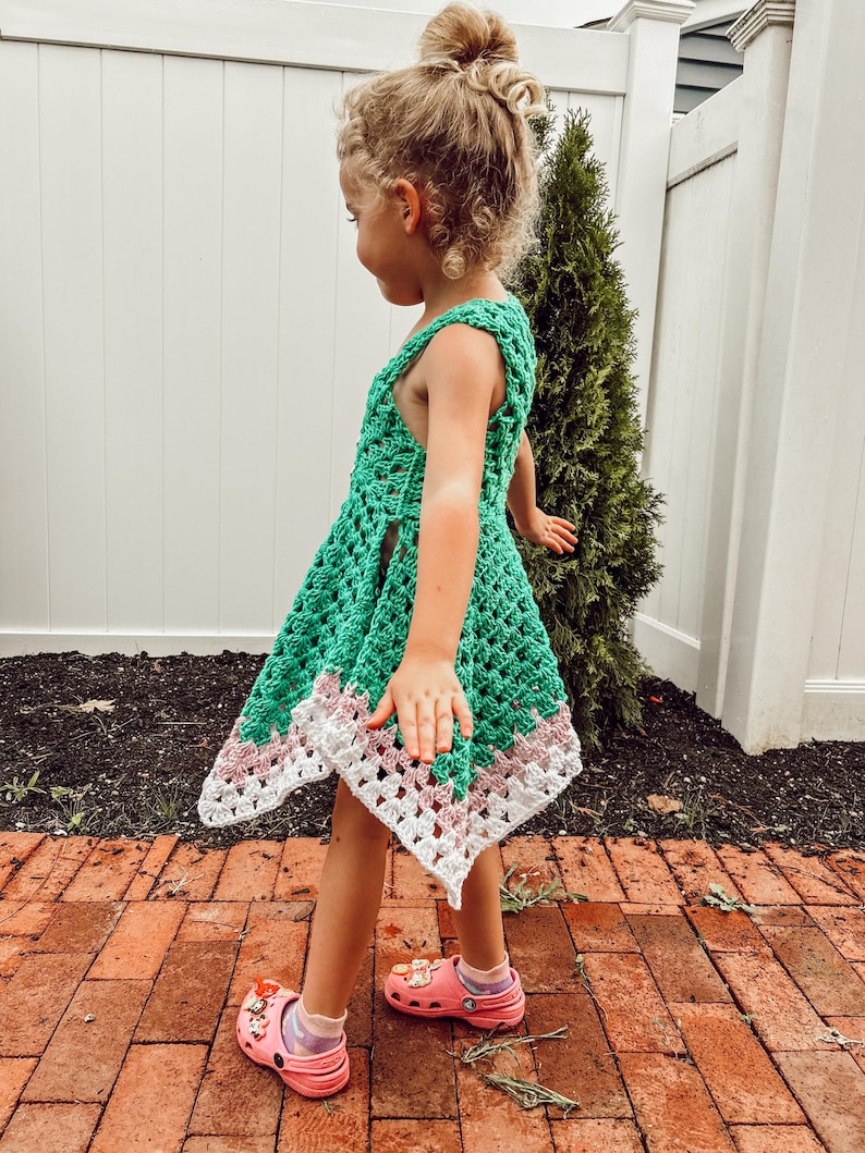 May include: A green crochet dress with a white and pink border. The dress is sleeveless and has a flared skirt. The child is wearing pink clogs.