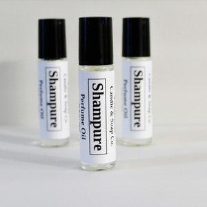 May include: Three clear glass roll-on perfume oil bottles with black caps. Each bottle has a white label with the text "Shampure Perfume Oil" and "Candle & Soap Co." in black. The bottles are arranged in a triangular pattern.
