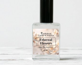 May include: A clear glass perfume bottle with a black cap. The label reads "Ethereal Flowers" and "Eau de Parfum 60 ML" over a floral background. The bottle is from "Fiddlehead Candle & Soap Co."