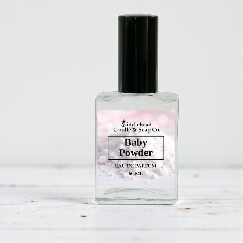 Może przedstawiać: Przezroczysty szklany flakon perfum z czarną nakrętką. Etykieta głosi "Baby Powder" i "Eau de Parfum 60 ml". Butelka pochodzi od "Fiddlehead Candle & Soap Co."