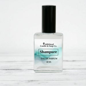 May include: A clear glass perfume bottle with a black cap. The label reads "Shampure Eau de Parfum 60 ML" with the "Fiddlehead Candle & Soap Co." logo. The background has a soft, blurred teal and white design.