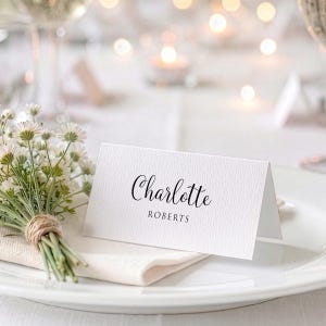 May include: A white place setting with a folded name card reading "Charlotte Roberts" in black script. A small bouquet of white flowers tied with twine sits on a napkin to the left of the card. Soft lighting and a blurred background.
