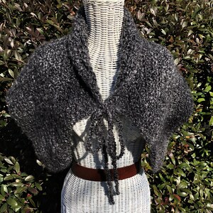 May include: A dark gray, hand-knitted shawl with a textured, open weave. The shawl has a draped design with a tie closure at the front. The garment is displayed on a white wicker form, with a brown belt visible below.
