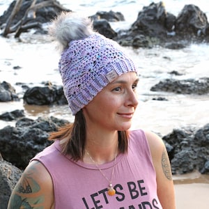 Puede incluir: Una mujer lleva una boina de ganchillo de color morado y blanco con un pompón esponjoso en la parte superior. La boina tiene una pequeña etiqueta en el lateral. Lleva una camiseta de tirantes rosa con el texto "Let's Be Mermaids" en ella. Está de pie frente a una playa rocosa.