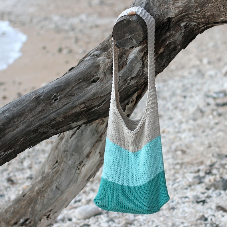 May include: A knitted tote bag with a beige top section and a teal and light blue bottom section. The bag is hanging from a branch.
