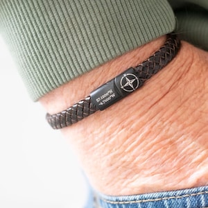 May include: A dark brown braided leather bracelet featuring a black metal clasp. The clasp is engraved with a compass rose symbol and coordinates that read 37.4220°N 122.0841°W. The bracelet is worn on a wrist partially tucked into a pocket.