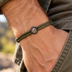 May include: A double-strand olive green cord bracelet featuring a small, circular black charm. The charm is engraved with a paw print graphic and the name "BUDDY" in white lettering, centered on the wrist with a simple adjustable design.