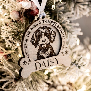 May include: Wooden dog ornament featuring a laser-etched portrait of a dog named Daisy. The bone-shaped ornament has the text "Deck the Paws with Bones of Holly" and snowflake details. It hangs from a white ribbon on a frosted Christmas tree.