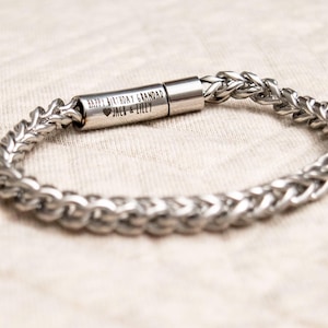 May include: A silver-toned metal chain bracelet featuring a cylindrical clasp engraved with the text HAPPY BIRTHDAY GRANDAD followed by a heart symbol and the names JACE & LILLY. The bracelet has a woven, braided link design.