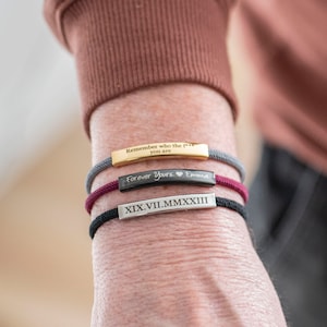 Op de afbeelding: Drie armbanden om een pols. De bovenste armband is grijs met de tekst "Remember who the f*** you are". De middelste armband is bordeauxrood met de tekst "Forever Yours" en een hartje. De onderste armband is zwart met de Romeinse cijfers "XIX VII.MMXXIII".
