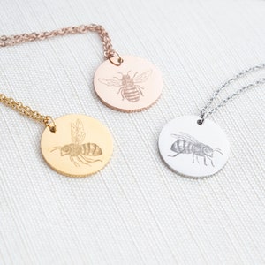 May include: Three necklaces in gold, rose gold, and silver with round pendants featuring an engraved bee design.