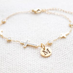 May include: Gold-coloured bracelet with cross and round charm details. The bracelet features multiple small crosses and clear, round accents. A small, round charm with the letter "M" hangs from the bracelet.