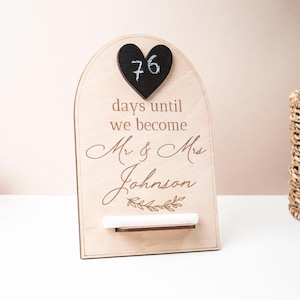 Könnte beinhalten: Ein hölzernes Hochzeits-Countdown-Schild mit einer schwarzen herzförmigen Tafel mit "76" Tagen. Auf dem Schild steht "Tage bis wir Mr. & Mrs. Johnson werden" mit floralen Akzenten. Eine silberne Vogelfigur und ein Weidenkorb sind ebenfalls zu sehen.