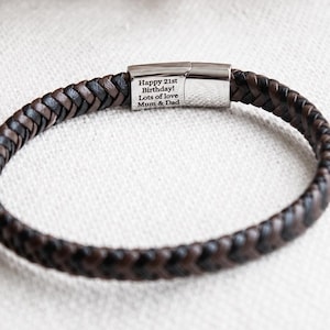 May include: A braided leather bracelet featuring dark brown and black strands. The bracelet includes a polished silver-tone metal magnetic clasp engraved with the text: Happy 21st Birthday! Lots of love Mom & Dad. The design is circular and simple.