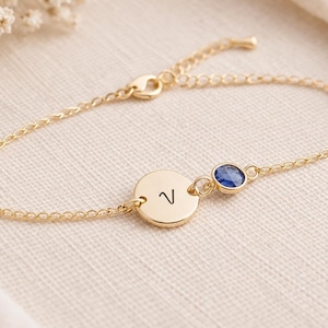 May include: A gold-coloured bracelet with a delicate chain. It features a round charm engraved with the letter "V", and a small blue gemstone. The bracelet is displayed on a cream-coloured fabric.
