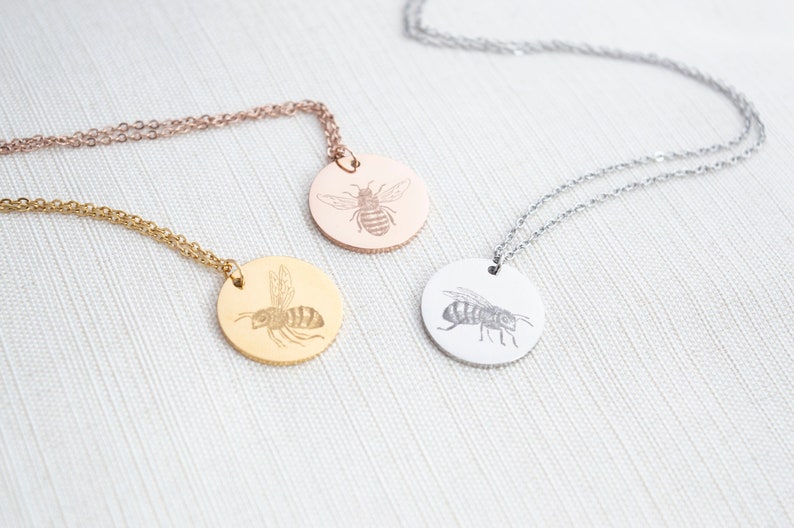 May include: Three necklaces in gold, rose gold, and silver with round pendants featuring an engraved bee design.
