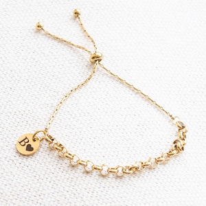Könnte beinhalten: Goldfarbenes Armband mit einer filigranen Kette und einem kleinen, runden Anhänger. Der Anhänger zeigt den Buchstaben "B" und ein Herzsymbol. Das Armband hat einen verstellbaren Schiebeknotenverschluss. Das Armband liegt auf weißem Untergrund.