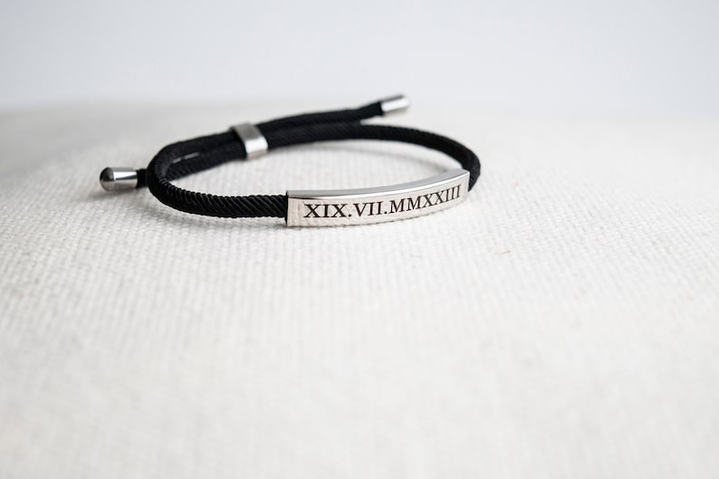A black braided cord bracelet with a silver rectangular tag engraved with the Roman numeral date "XIX.VII.MMXXIII".