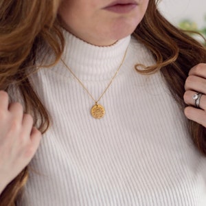 May include: A gold necklace with a round pendant featuring a detailed design. The necklace is worn by a person with long brown hair and a white turtleneck sweater.