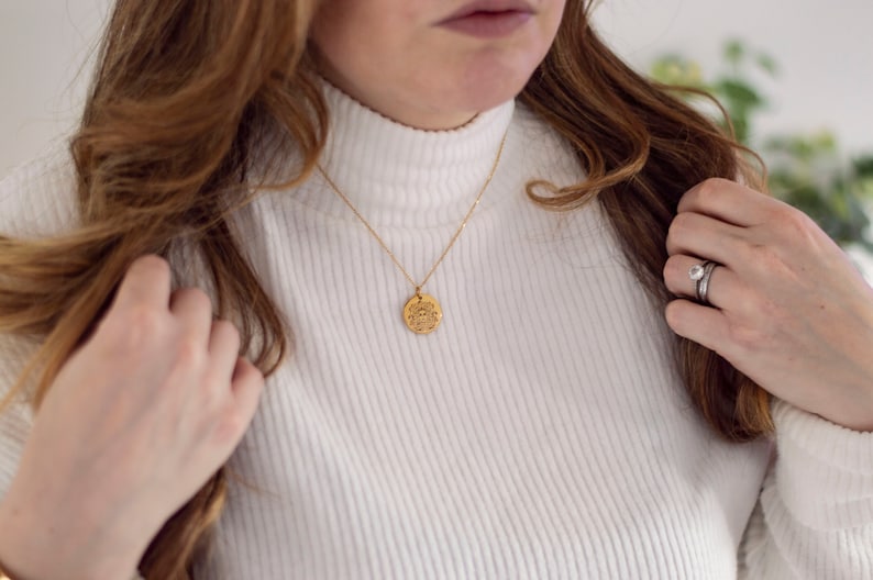 May include: A gold necklace with a round pendant featuring a detailed design. The necklace is worn by a person with long brown hair and a white turtleneck sweater.