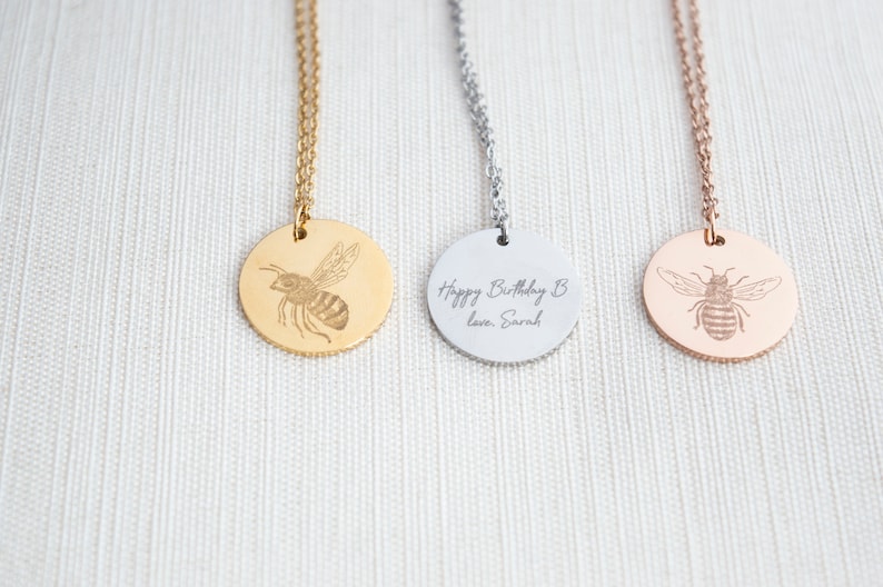 May include: Three gold, silver, and rose gold necklaces with round pendants featuring an engraved bee design. The silver pendant has the text "Happy Birthday B love, Sarah" engraved on it.
