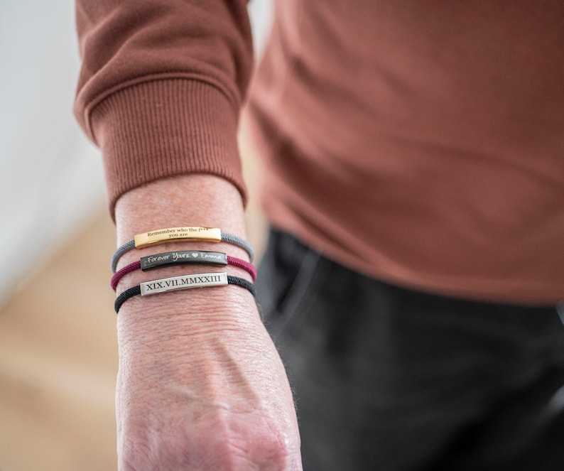 Op de afbeelding: Drie armbanden om een pols. De bovenste armband is grijs met de tekst "Remember who the f*** you are". De middelste armband is bordeauxrood met de tekst "Forever Yours" en een hartje. De onderste armband is zwart met de Romeinse cijfers "XIX VII.MMXXIII".