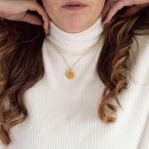 May include: A gold chain necklace with a round coin pendant. The coin has a detailed design and is worn by a person with long brown hair.