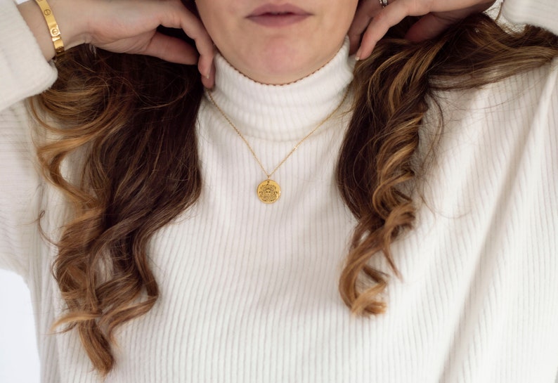 May include: A gold chain necklace with a round coin pendant. The coin has a detailed design and is worn by a person with long brown hair.