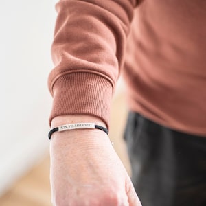 Op de afbeelding: Een zwart koordarmband met een zilveren staafbedel gegraveerd met Romeinse cijfers. De armband wordt om de pols gedragen.