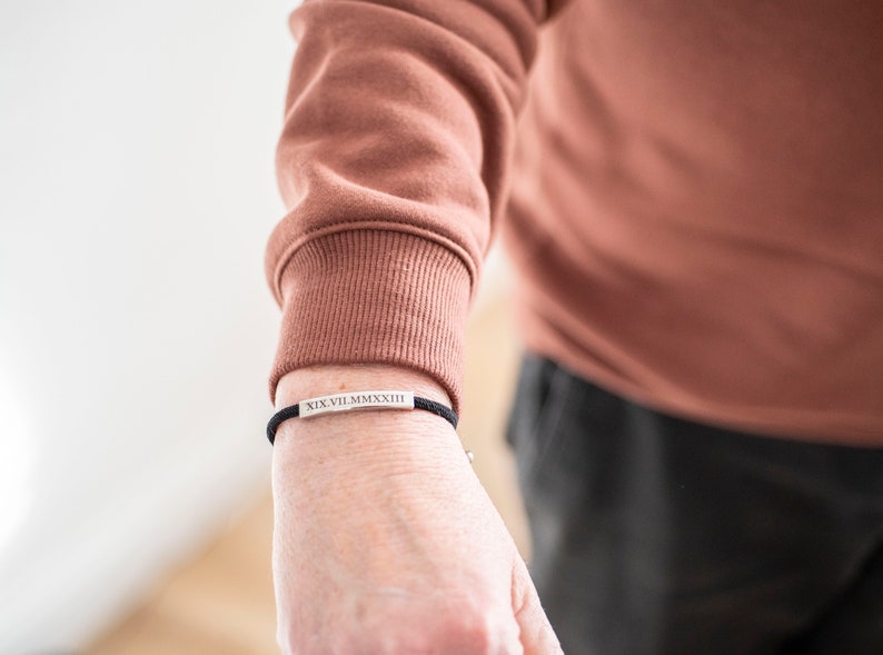Op de afbeelding: Een zwart koordarmband met een zilveren staafbedel gegraveerd met Romeinse cijfers. De armband wordt om de pols gedragen.