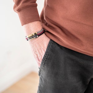 Op de afbeelding: Een persoon draagt een bruine trui en een zwarte broek met drie armbanden om de pols. De armbanden zijn zwart, bordeauxrood en goud.