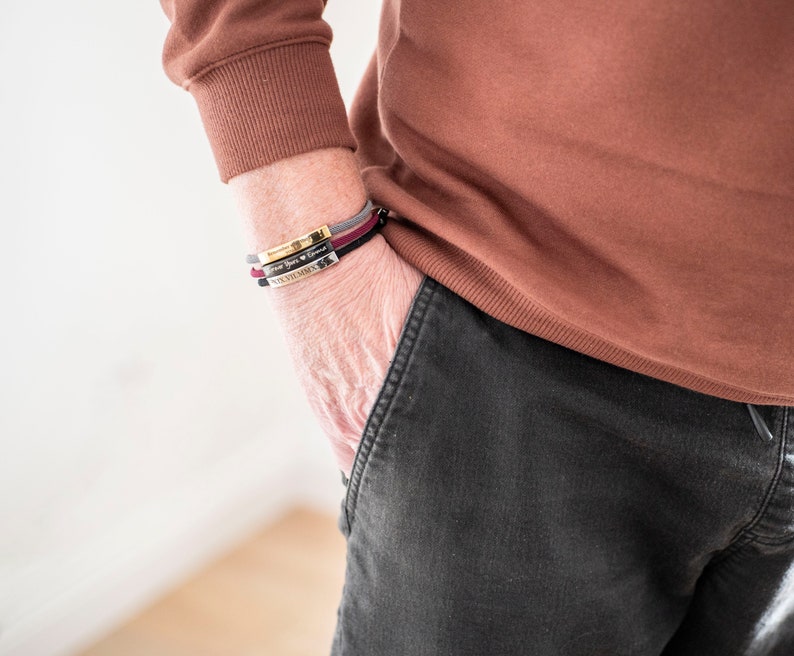 Op de afbeelding: Een persoon draagt een bruine trui en een zwarte broek met drie armbanden om de pols. De armbanden zijn zwart, bordeauxrood en goud.