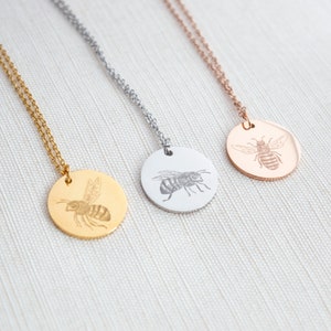 May include: Three gold, silver, and rose gold necklaces with a round pendant featuring an engraved bee design.