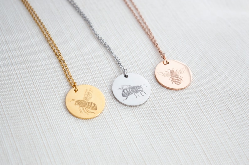 May include: Three gold, silver, and rose gold necklaces with a round pendant featuring an engraved bee design.