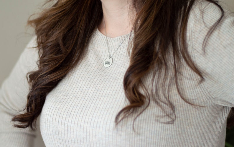 May include: A woman wearing a light beige ribbed sweater with a silver chain necklace and a small oval pendant.