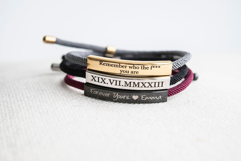 Op de afbeelding: Drie armbanden met gegraveerde metalen platen. De bovenste plaat is goudkleurig en heeft de tekst "Remember who the f*** you are". De middelste plaat is zilverkleurig en heeft de tekst "XIX.VII.MMXXIII". De onderste plaat is zwart en heeft de tekst "Forever Yours. &hearts; Emma".