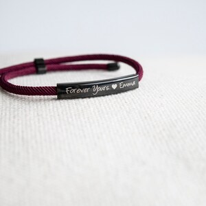 Op de afbeelding: Burgundy rood gevlochten koord armband met een zwarte metalen balk gegraveerd met de tekst "Forever Yours. &hearts; Emma"