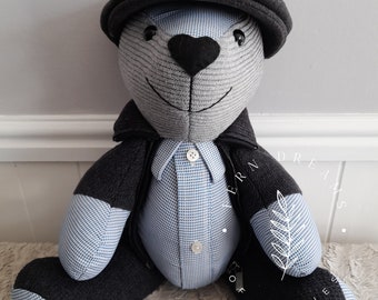 May include: A grey and blue teddy bear wearing a black jacket, a blue and white checked shirt, and a grey cap. The bear has black eyes and a black nose. The bear's paws have the words "Love you Dad" and "Dream of Fairies" embroidered on them.