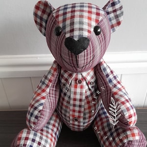 May include: A red, white and blue plaid teddy bear with black button eyes and a black heart-shaped nose. The bear is sitting on a dark brown surface. The bear is made from repurposed fabric.