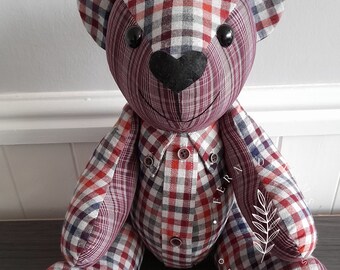 May include: A red, white, and blue plaid teddy bear with black button eyes and a black heart-shaped nose. The bear is sitting on a dark brown surface. The bear is made from repurposed fabric.