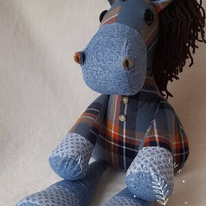 May include: A handmade plush horse doll made from blue denim and plaid fabric. The horse features brown yarn hair, button eyes, and the words "Dad" and "God Bless" printed on its feet. The doll is positioned on a white surface.